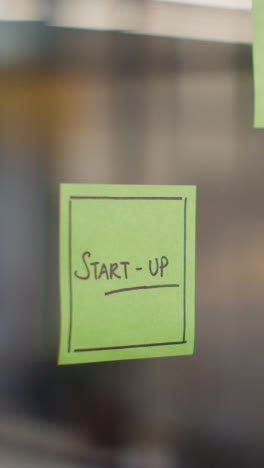 Vertical-Video-Close-Up-Of-Woman-Putting-Sticky-Note-With-Start-Up-Written-On-It-Onto-Transparent-Screen-In-Office